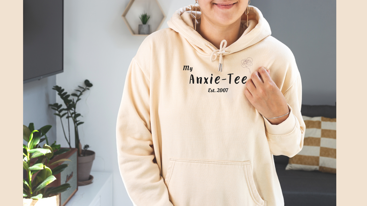 person wearing a cream colored hoodie with the words My Anxie-Tee Est. 2007 across the front.